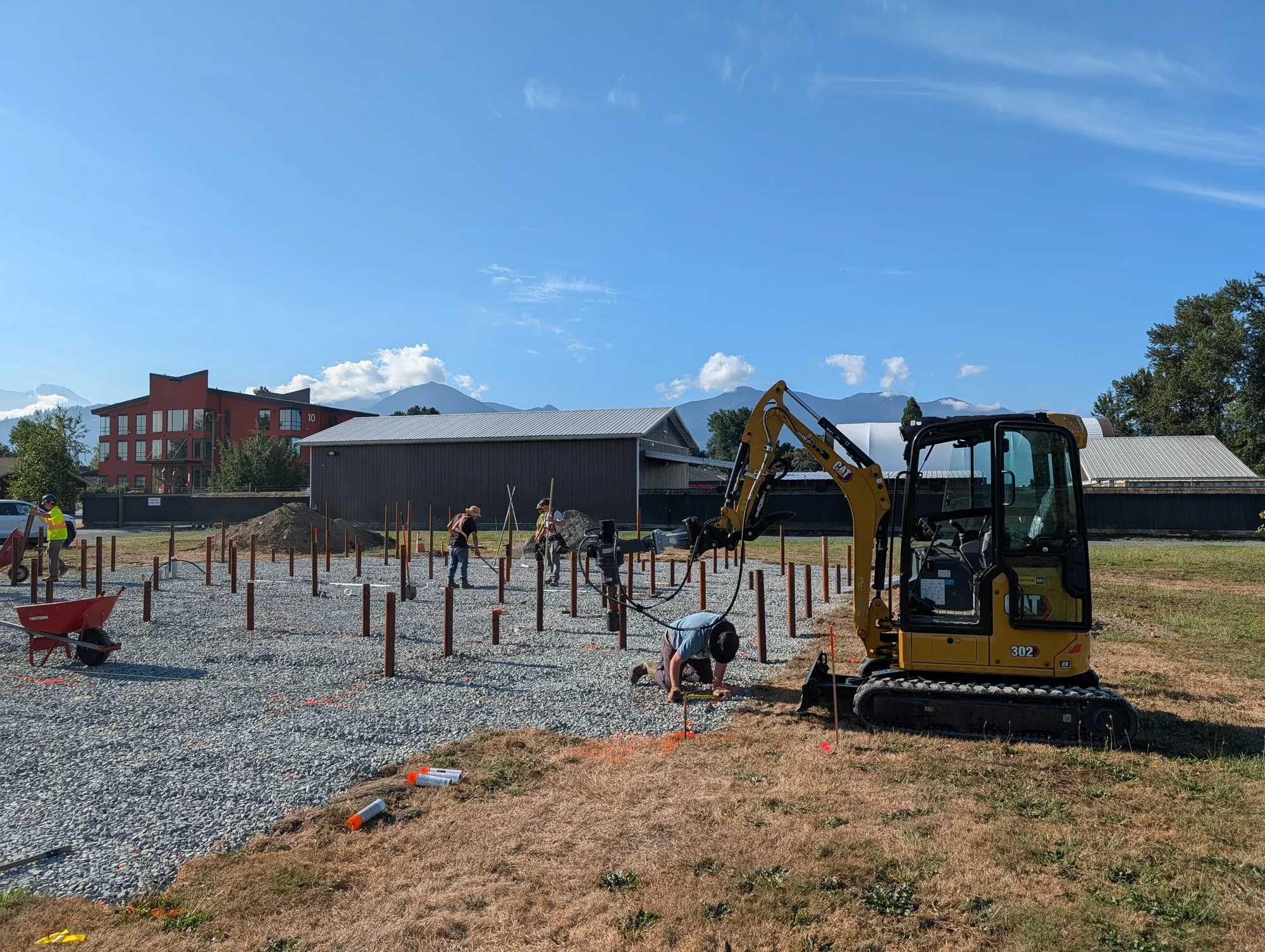Office Building - Compact excavator driving piles across the pad while the crew checks each position